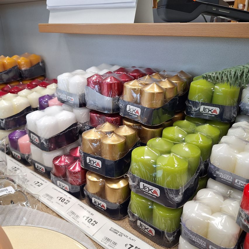 A store shelf displays an assortment of Jeka candles, including various colors like white, green, red, and gold, all individually wrapped. The display is neat with labels and price tags visible.
