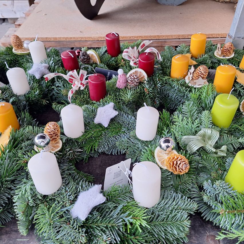 Ein kreisförmiger Kranz aus Tannenzweigen, mit verschiedenfarbigen Kerzen und Dekorationen, einschließlich Tannenzapfen und Bändern, auf einem Tisch ausgestellt.