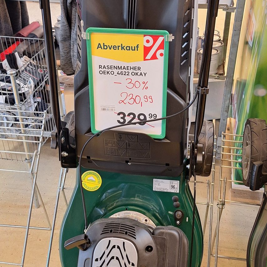 A green lawn mower displays a discount sign with the price of 230.99 euros and a 30% reduction. It is located in a retail store.