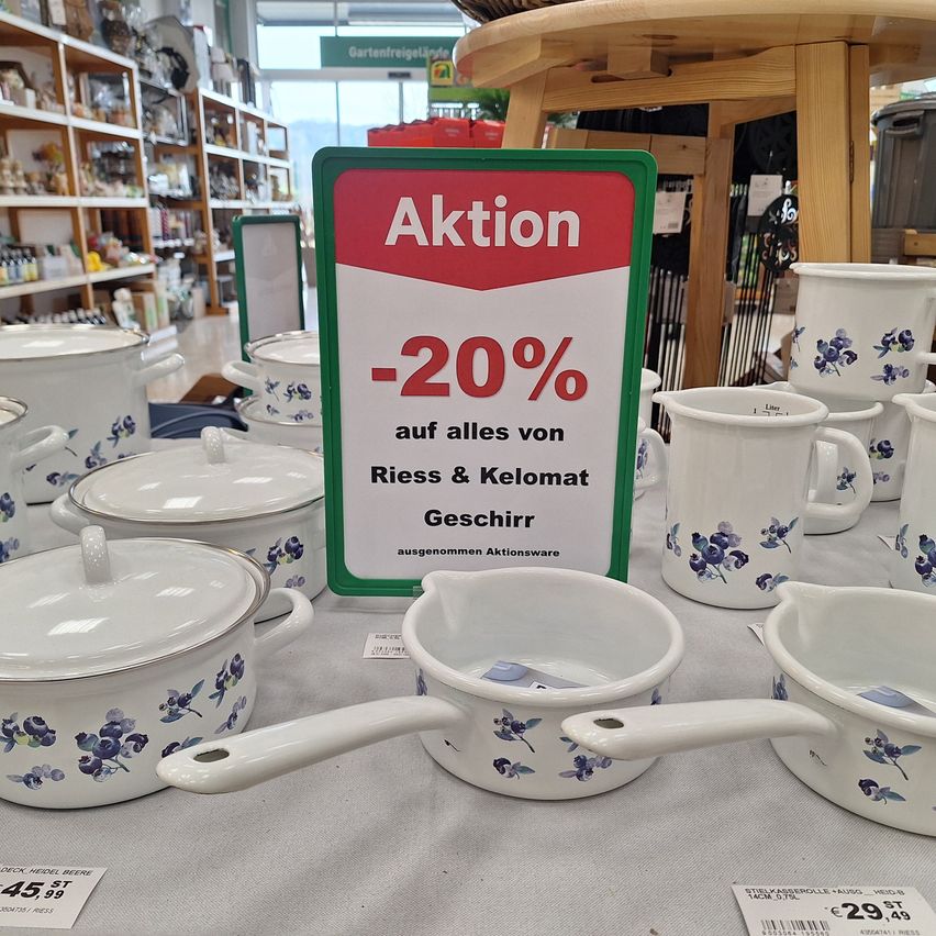 Ein Tischdisplay mit verschiedenen Keramiktöpfen und -pfannen mit blauen Blumendesigns. Ein Schild mit 'Aktion -20%' in Rot und Weiß. Die Produkte stammen von Riess & Kelomat.