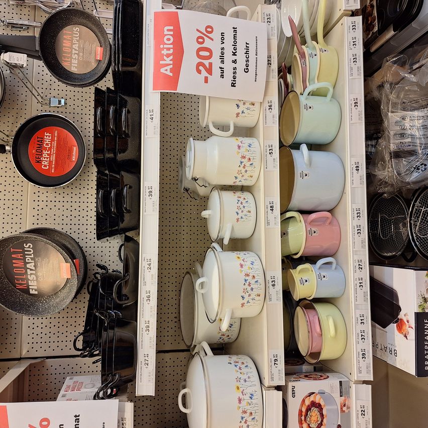 Ein Ladenregal zeigt verschiedene Kochtöpfe und Pfannen, darunter ein weißer Topf mit Blumendesigns. Ein Schild weist auf einen 20%igen Rabatt auf alle Artikel hin. Schwarze Bratpfannen sind ebenfalls an der Wand zu sehen.