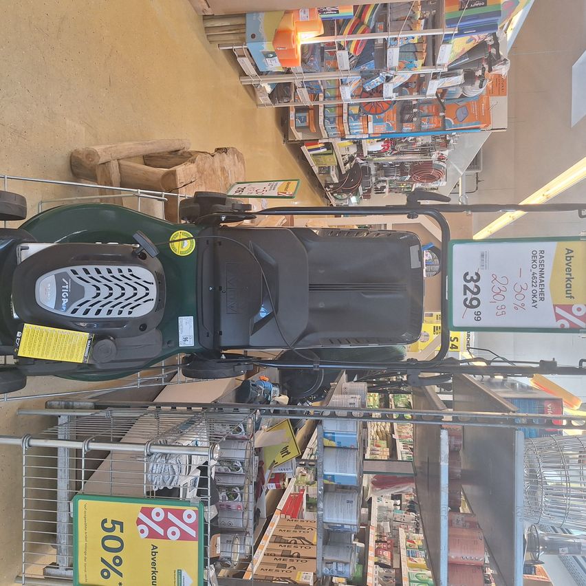 A green lawn mower is on display in a store with a price tag of 329.99 euros. The store has a sale sign with a 50% discount.