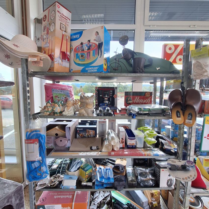 A cluttered shelf displays various household items, including toys, cleaning supplies, and a pair of slippers. The background shows a store with a glass window and a discount sign.