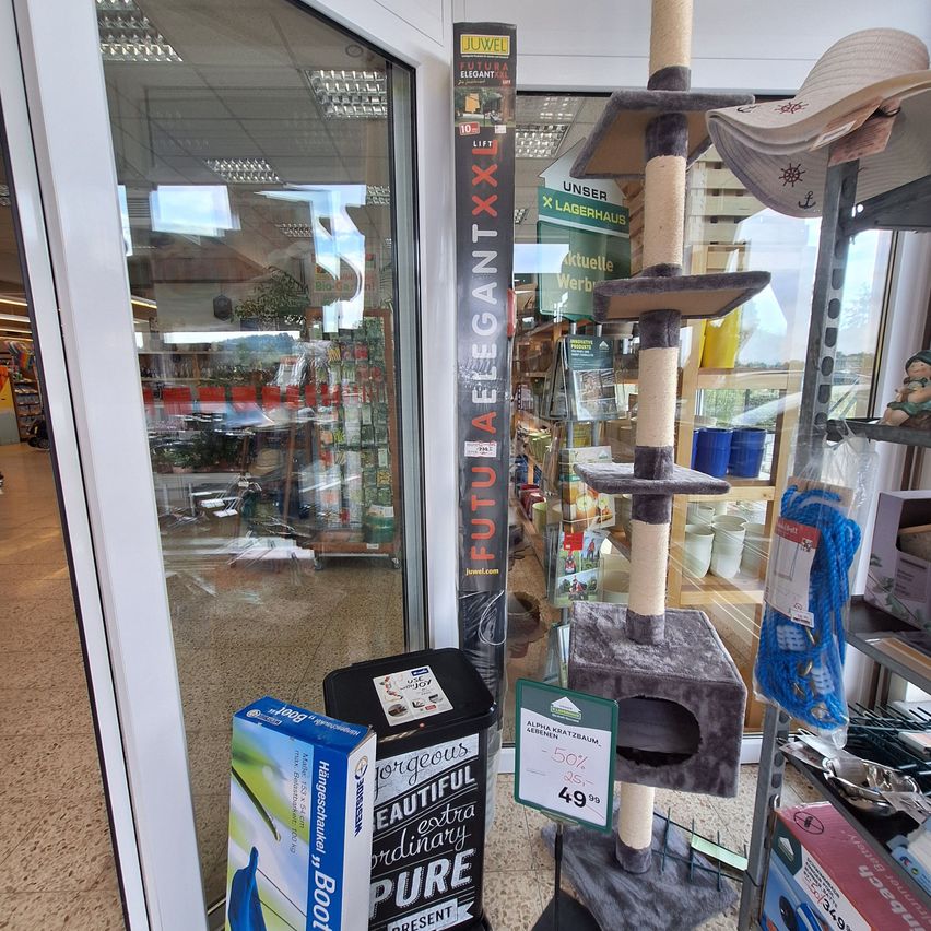 A cat tree is displayed in a store with a sign that reads 'FUTURELEGANT XXL'. A trash bin and a box are placed next to the cat tree. The store has glass walls and shelves with various items.