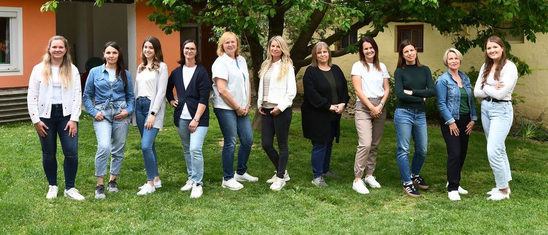 Eine Gruppe von Frauen posiert für ein Foto vor einem großen Baum, der auf einem Rasen steht. Sie sind alle lässig gekleidet und tragen Turnschuhe. Der Baum ist üppig mit grünen Blättern. Hinter ihnen steht ein Gebäude mit einer orangefarbenen Wand und einem Fenster.