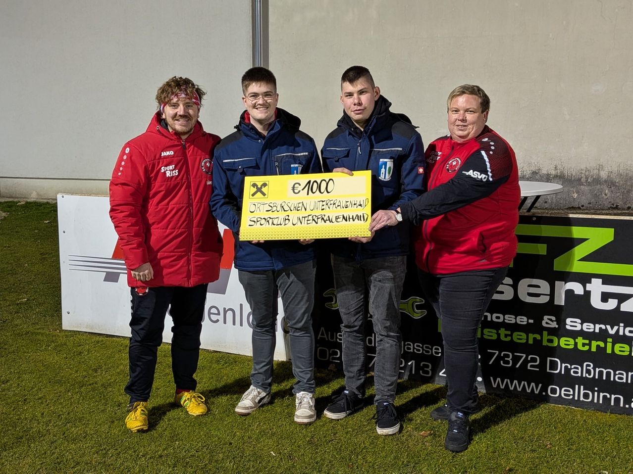 Vier Personen stehen auf einem Rasenfeld und halten ein großes gelbes Banner mit einem Preis von 1000 Euro. Das Banner wird von zwei Männern in blauen Jacken gehalten. Dahinter lächelt ein Mann in einer roten Jacke. Ein Mann in einer roten und schwarzen Jacke steht neben ihnen.
