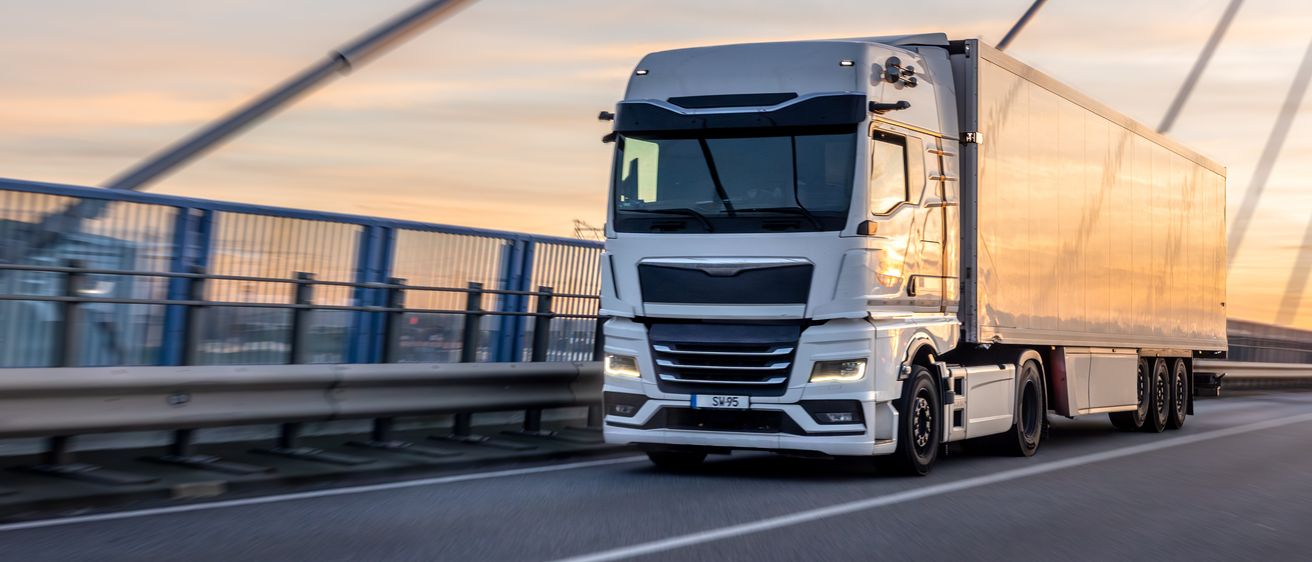 Ein weißer Sattelschlepper fährt bei Dämmerung auf einer Brücke. Der LKW hat einen großen Laderaum und ist beleuchtet.