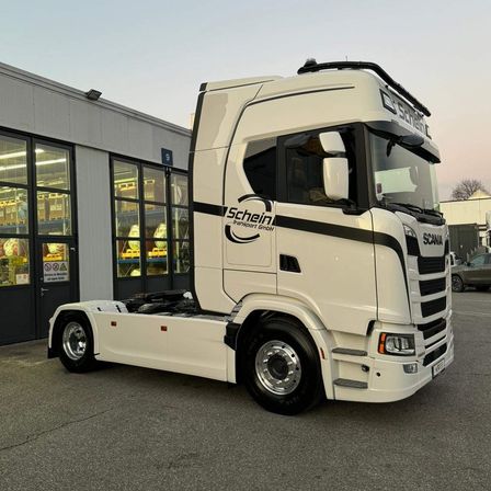 Ein weißer Scania-Lkw mit schwarzen Streifen ist vor einem Gebäude mit Glaswänden geparkt, in der Nähe ist ein Auto geparkt.