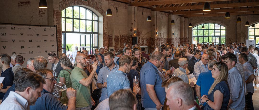 Eine große Gruppe von Menschen in einem großen Raum trinkt Wein. Sie stehen und unterhalten sich miteinander. Der Raum hat Holzbalken und Ziegelwände.