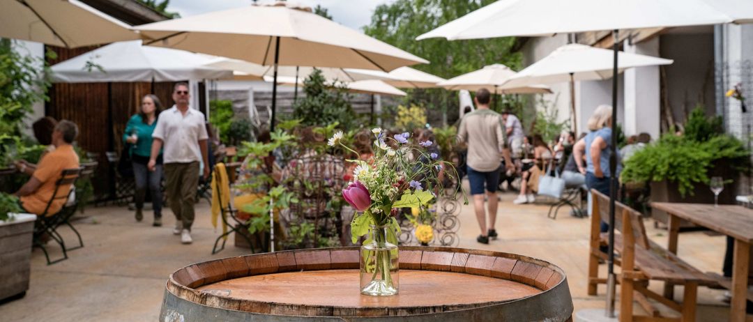 Eine Außengastronomie mit weißen Sonnenschirmen und einem Holzfass mit Blumen. Leute gehen auf dem Pfad.