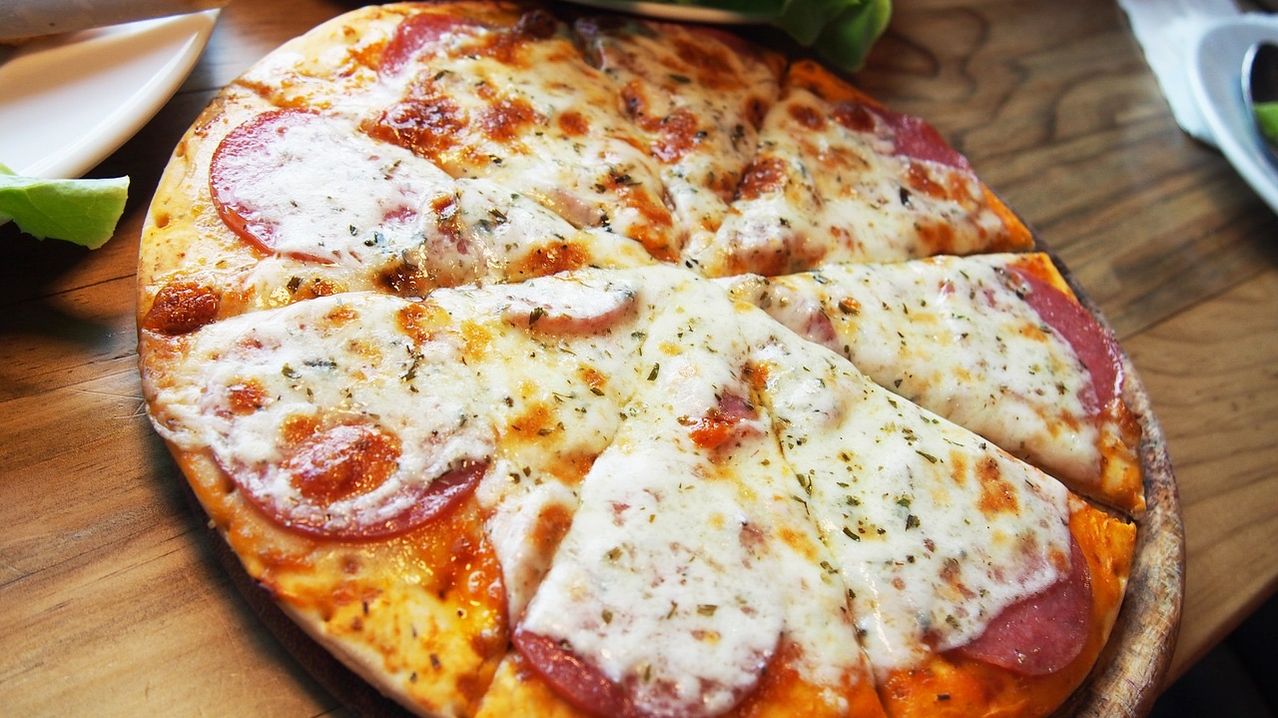 Eine frisch gebackene Pizza mit einer dicken Käseschicht, belegt mit Peperoni und Kräutern. Sie ist in acht Stücke geschnitten auf einer Holzoberfläche.