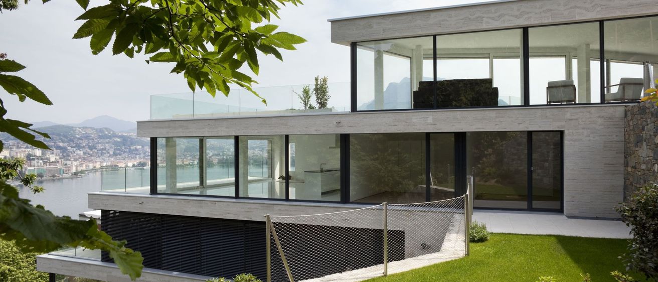 Modernes Haus mit Glaswänden und Balkon am Hang mit Blick auf das Wasser.