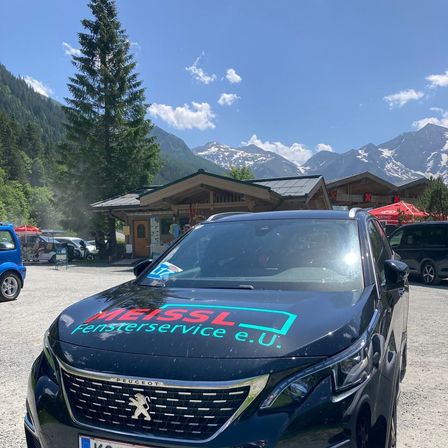 Ein Peugeot-Auto mit dem Text 'MEISSEL Fensterservice e.U.' geparkt vor einem Chalet mit Bergen im Hintergrund.