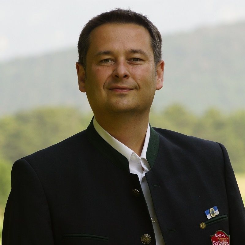 Ein Mann in einem traditionellen deutschen Anzug steht auf einem Feld mit Bergen im Hintergrund. Er trägt ein weißes Hemd und einen schwarzen Anzug mit grünem Besatz und verschiedenen Abzeichen auf seiner Jacke.