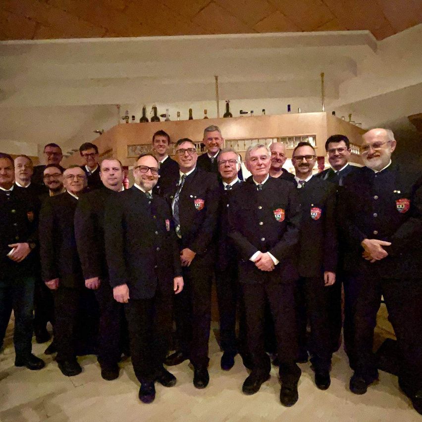 Eine Gruppe von Männern in formeller Kleidung, wahrscheinlich Feuerwehrleute, posiert für ein Foto in einem gut beleuchteten Raum mit einer Holzdecke.