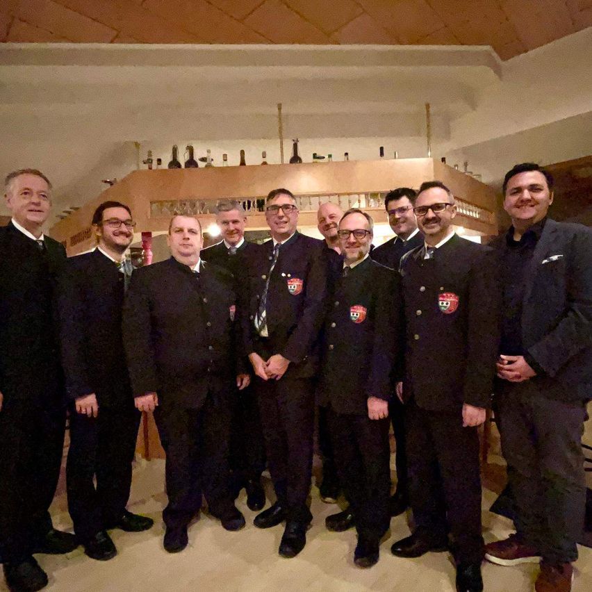 Eine Gruppe von Männern in formeller Kleidung, die Uniformen tragen, stehen zusammen in einem Raum, wahrscheinlich für ein Gruppenfoto. Sie alle lächeln. Hinter ihnen befindet sich ein Regal mit Flaschen.