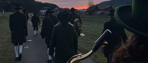 Eine Gruppe von Musikern in traditioneller Kleidung geht bei Sonnenuntergang einen Pfad entlang und spielt Blechblasinstrumente.