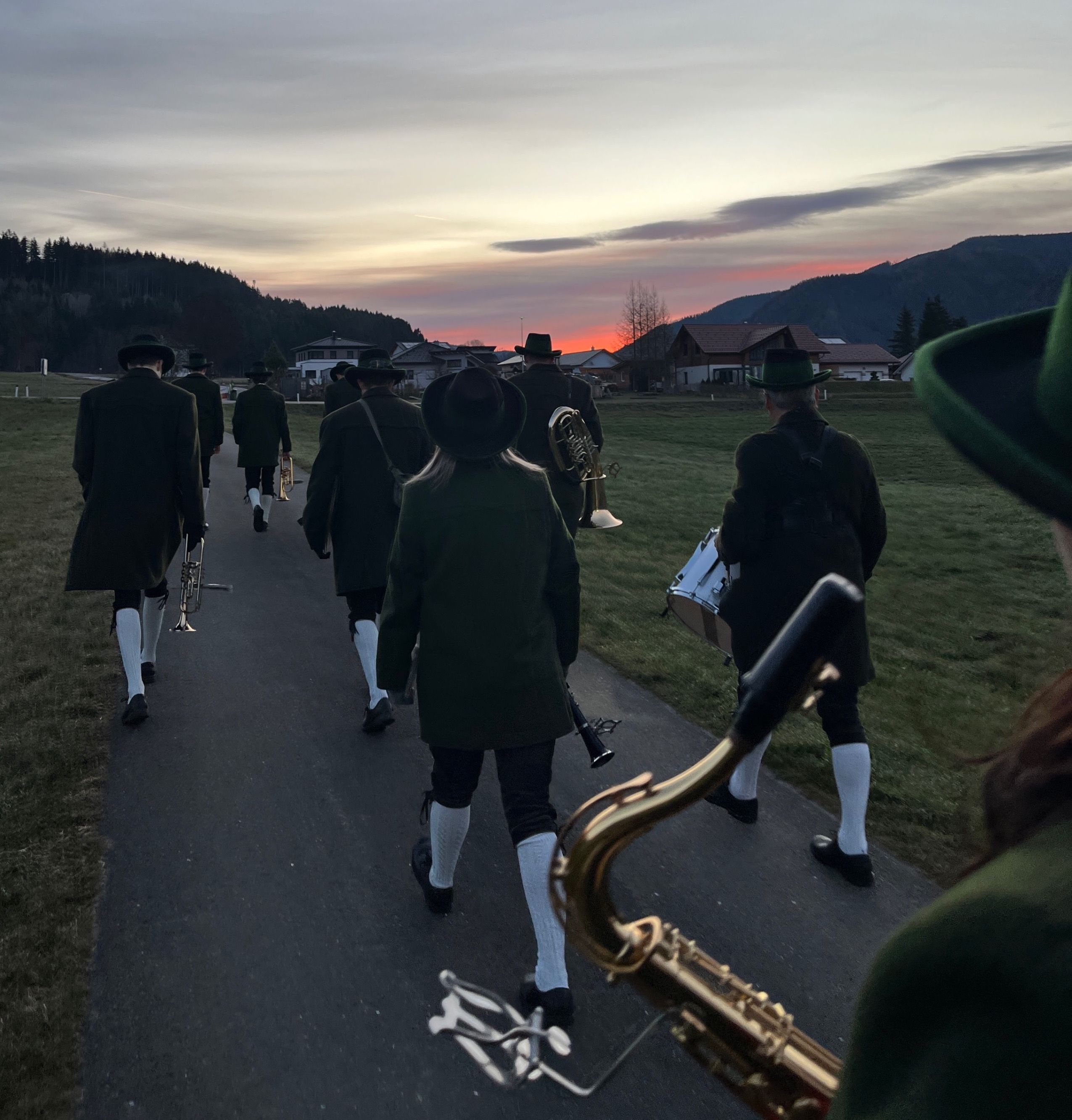 Eine Gruppe von Musikern in traditioneller Kleidung geht bei Sonnenuntergang einen Pfad entlang und trägt verschiedene Instrumente. Der Himmel ist in Orange- und Rosatöne getaucht.