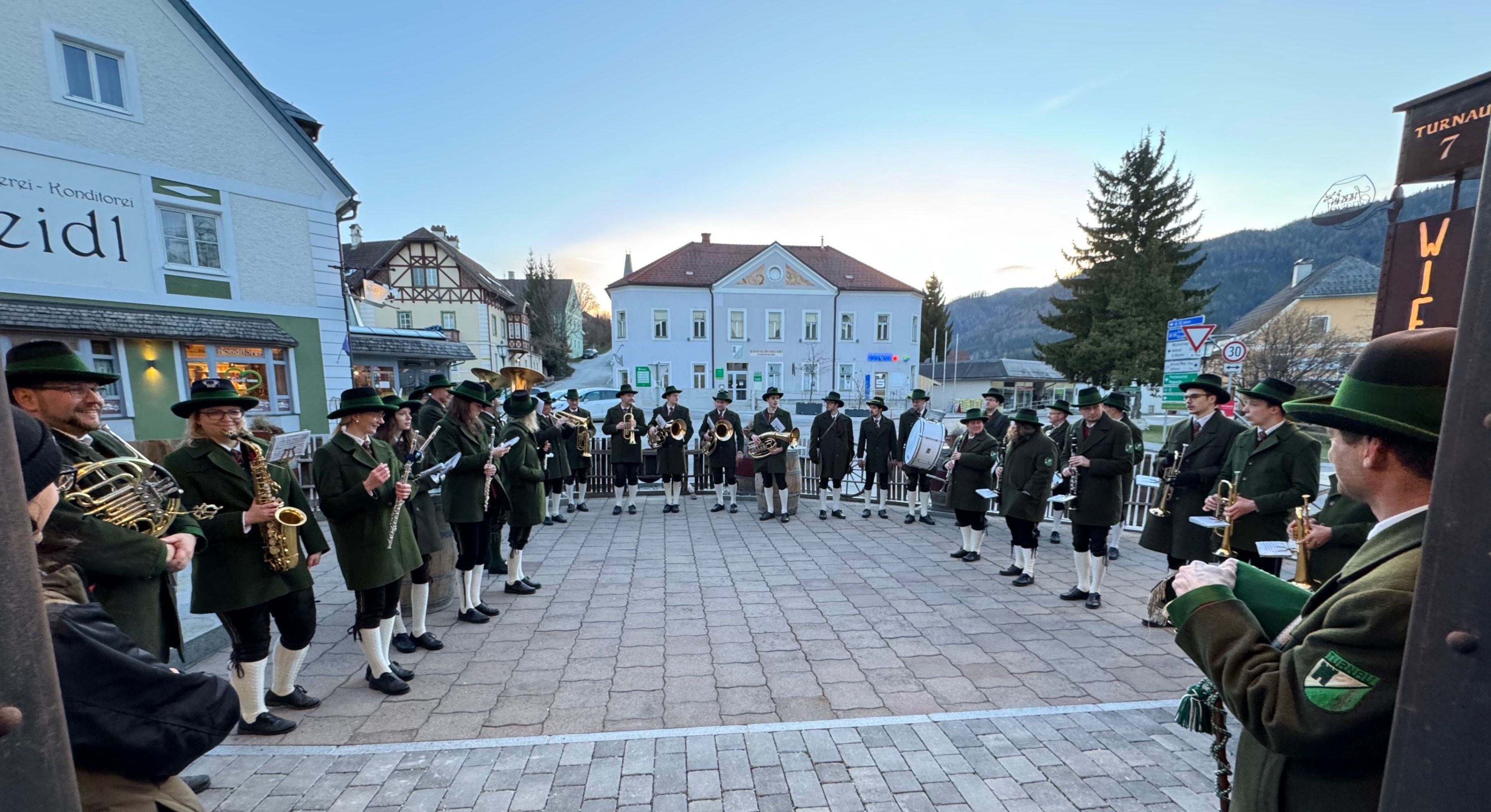 Eine Gruppe von Musikern in traditionellen grünen Uniformen und Hüten spielt im Freien vor einem Gebäude. Sie halten verschiedene Musikinstrumente wie Trompeten, Posaunen und Trommeln. Die Umgebung ist tagsüber mit klarem Himmel.