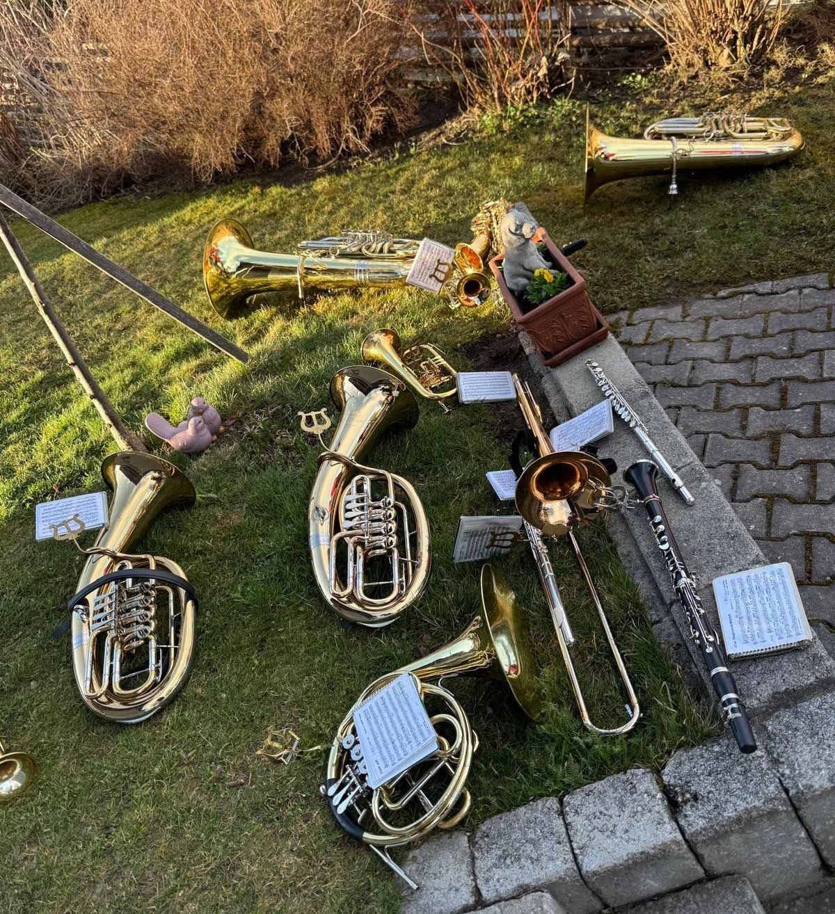 Eine Sammlung von Blech- und Holzblasinstrumenten liegt auf einem Grasbereich mit einer Ziegelsteinkante. Einige Instrumente haben Musikblätter, die an ihnen befestigt sind.