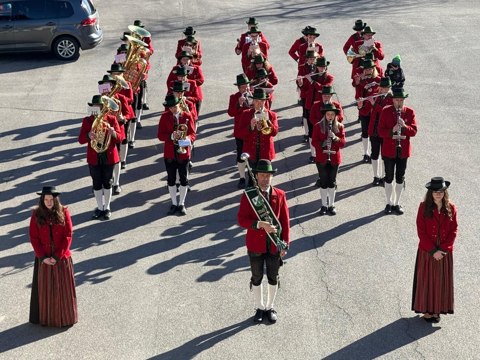 Eine Marschkapelle in roten Uniformen und grünen Hüten, mit verschiedenen Instrumenten, steht in Formation auf einer gepflasterten Straße.