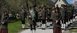 Eine Blaskapelle in grünen Uniformen und mit Musikinstrumenten steht in einer Reihe auf einer Straße, mit einer Frau an der Seite. Im Hintergrund befindet sich eine Bergkette mit Bäumen und einem Haus.