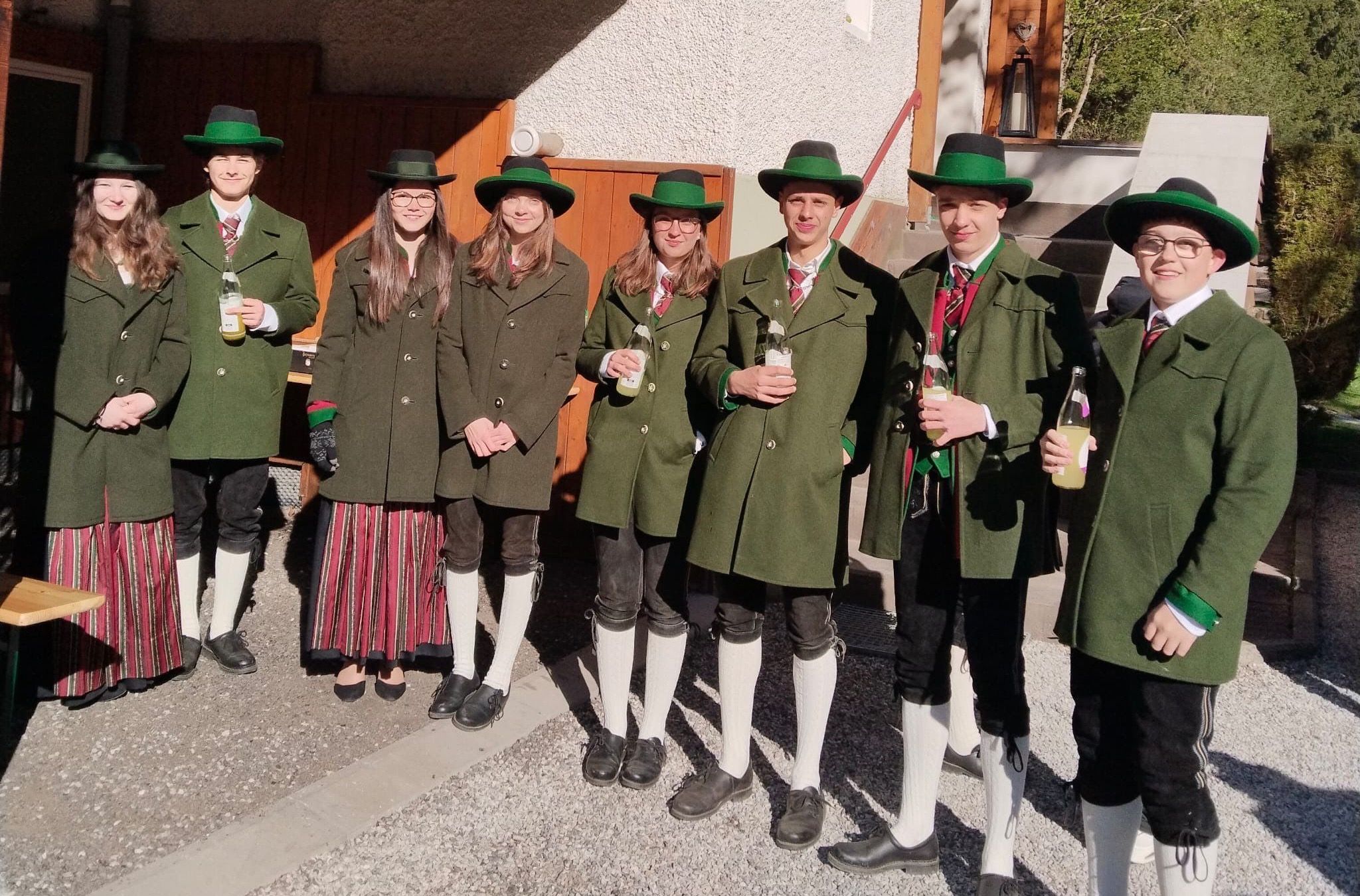 Eine Gruppe junger Menschen in grünen Mänteln und Hüten steht draußen und hält Flaschen. Sie scheinen für ein Foto zu posieren.