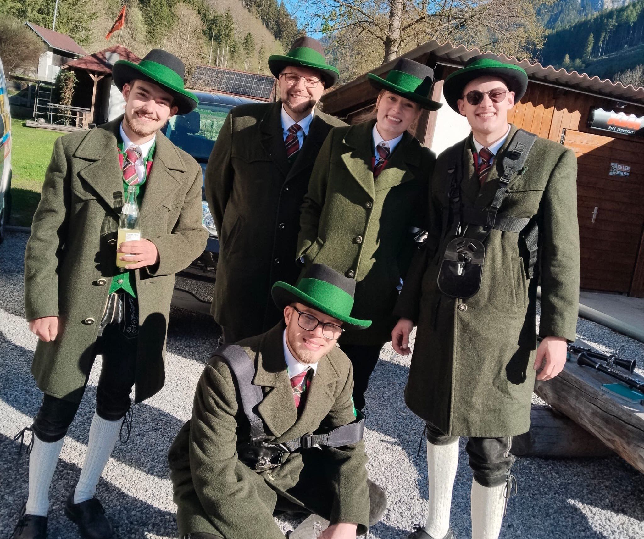 Fünf Personen, die in traditionellen grünen Mänteln und Hüten gekleidet sind, zwei davon kniend, posieren für ein Foto im Freien mit einem Berg im Hintergrund.