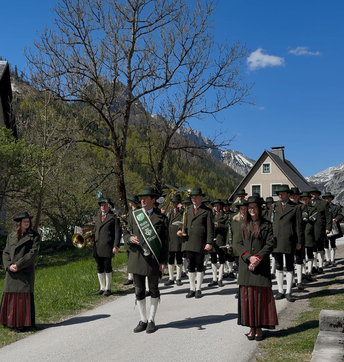 Ein Marschkapelle in traditioneller bayerischer Kleidung, einschließlich grüner Mäntel und weißer Socken, tritt an einem sonnigen Tag in einer Bergregion auf. Die Kapelle wird von einer Person angeführt, die eine grüne Fahne trägt.