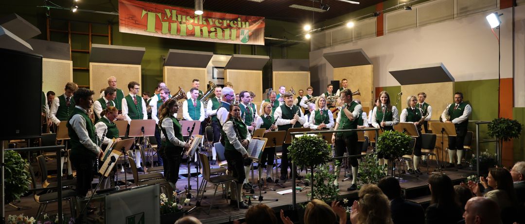 Eine Blaskapelle spielt auf der Bühne in grünen Westen, umgeben von Topfpflanzen und einem applaudierenden Publikum. Die Bühne hat ein Banner mit der Aufschrift 'Musikverein Turnau'.