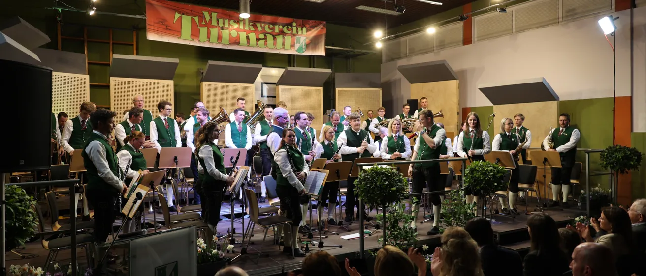 Eine Blaskapelle spielt auf der Bühne in grünen Westen, umgeben von Topfpflanzen und einem applaudierenden Publikum. Die Bühne hat ein Banner mit der Aufschrift 'Musikverein Turnau'.