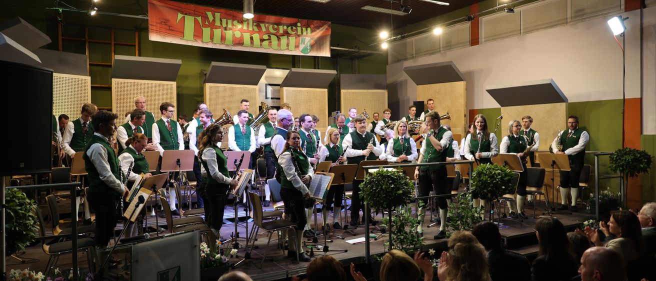 Eine Blaskapelle spielt auf der Bühne in grünen Westen, umgeben von Topfpflanzen und einem applaudierenden Publikum. Die Bühne hat ein Banner mit der Aufschrift 'Musikverein Turnau'.