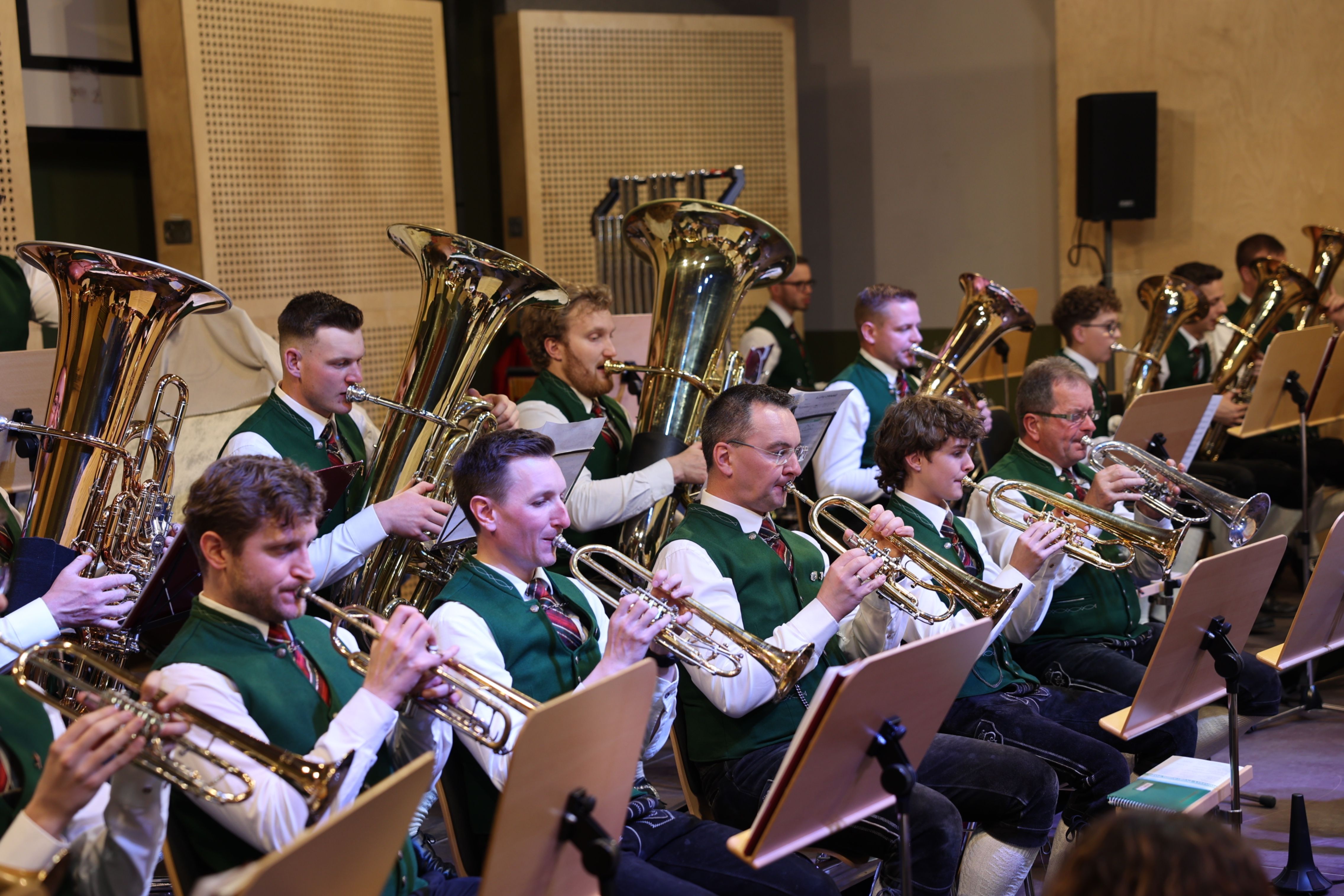 Eine Gruppe von Musikern in grünen Westen und weißen Langarmhemden spielt Blechblasinstrumente, darunter Trompeten und Tubas, in einer gut beleuchteten Innenumgebung.