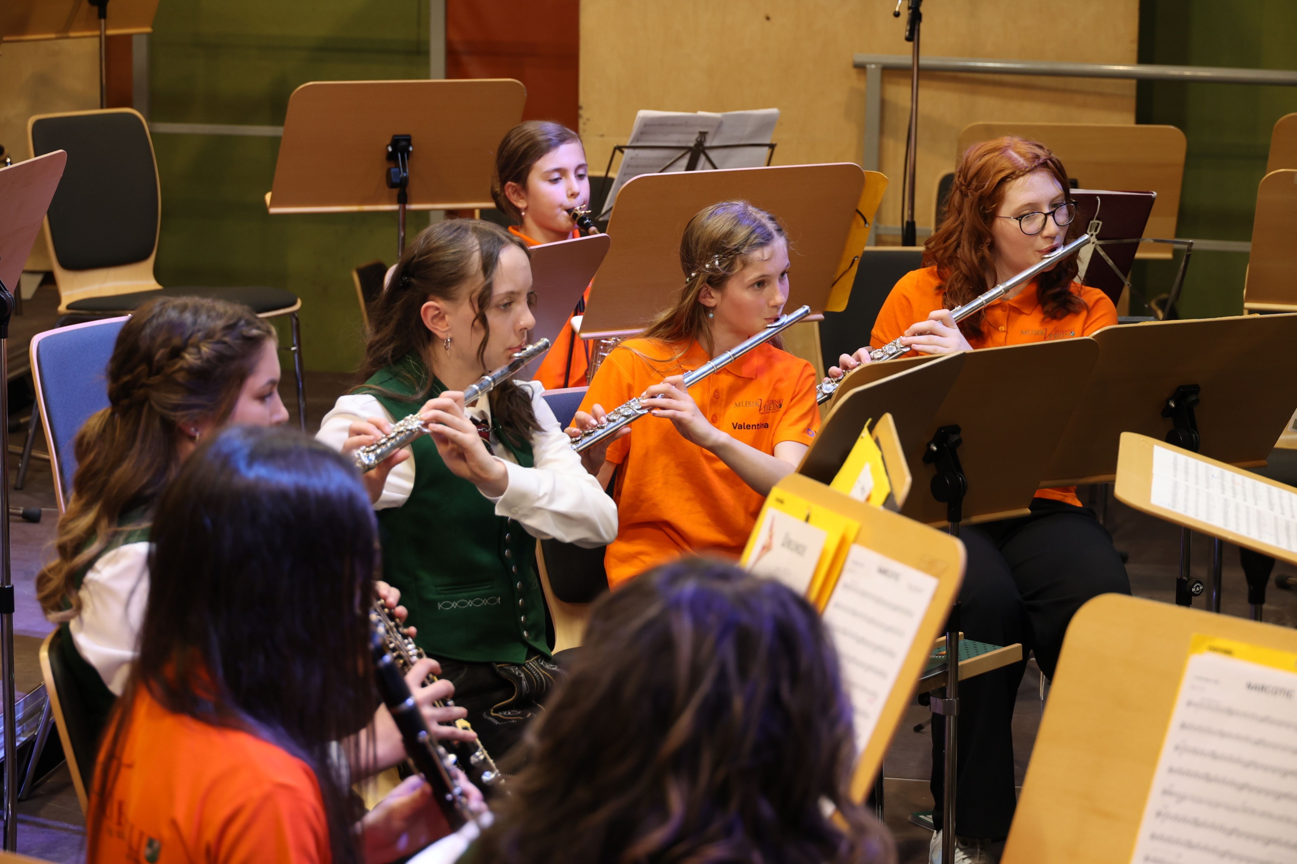 Eine Gruppe junger Mädchen in orangefarbenen Shirts spielt Flöten in einem Musikraum. Sie sitzen und konzentrieren sich auf ihre Instrumente.