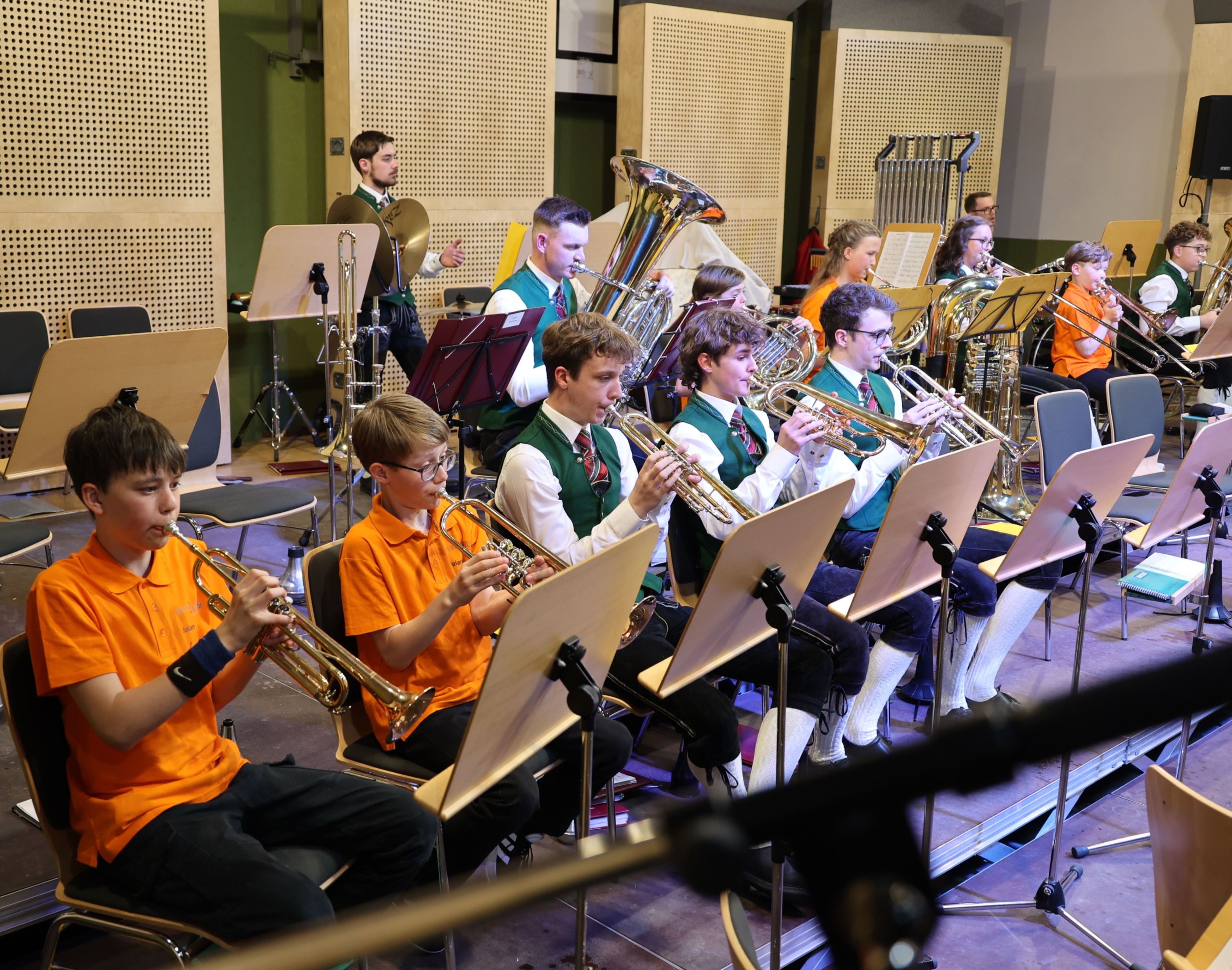 Eine Gruppe junger Musiker in grünen Westen und orangefarbenen Hemden spielt Trompeten in einem gut beleuchteten Proberaum. Sie sitzen vor Notenständern mit verschiedenen Instrumenten.
