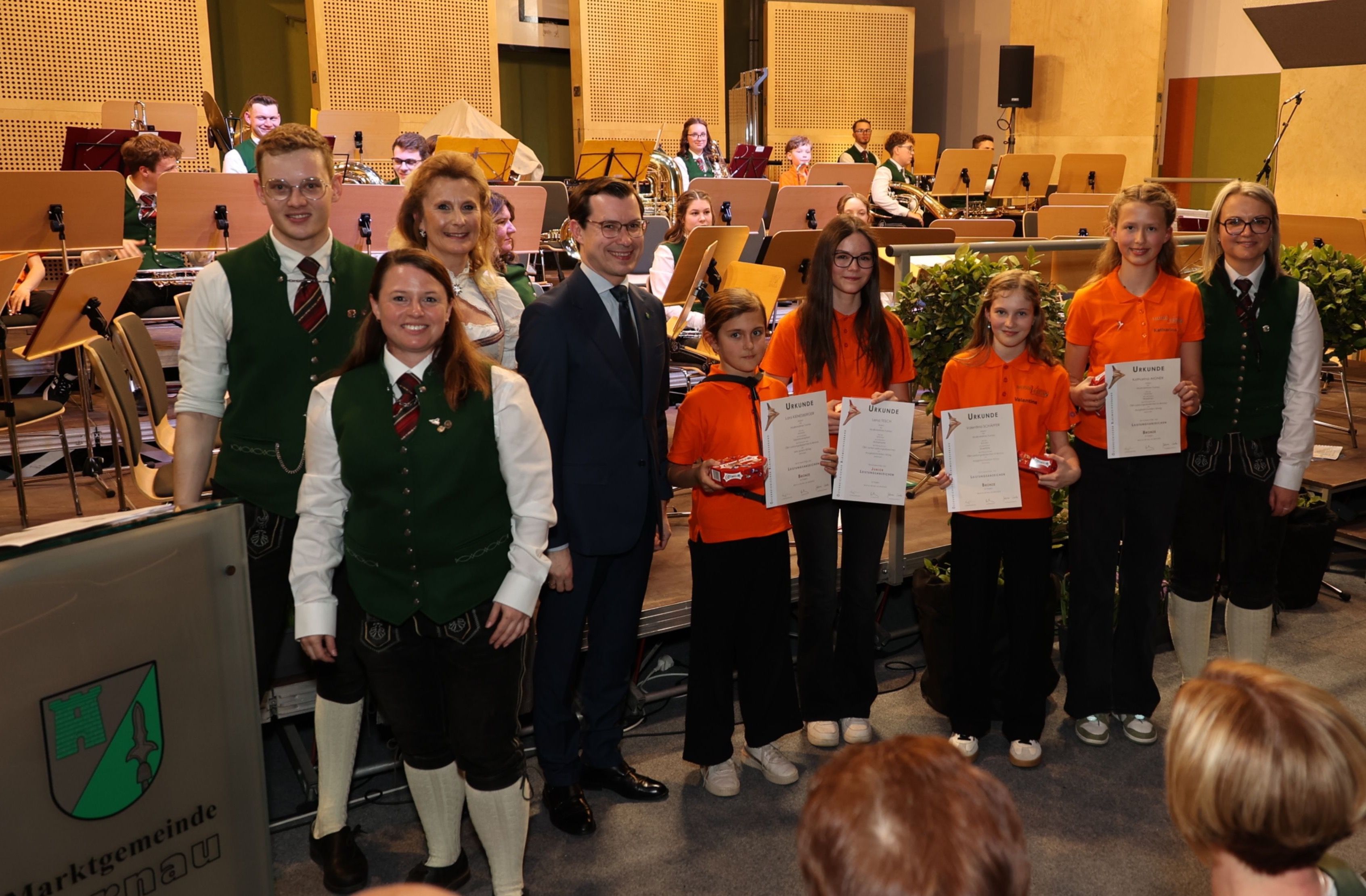 Eine Gruppe von Menschen in orangefarbenen Shirts und schwarzen Hosen steht auf einer Bühne mit einem Mann in Anzug und einer Frau in einem grünen West. Sie halten Urkunden und eine Schachtel mit Süßigkeiten. Hinter ihnen spielt eine Band mit Instrumenten.
