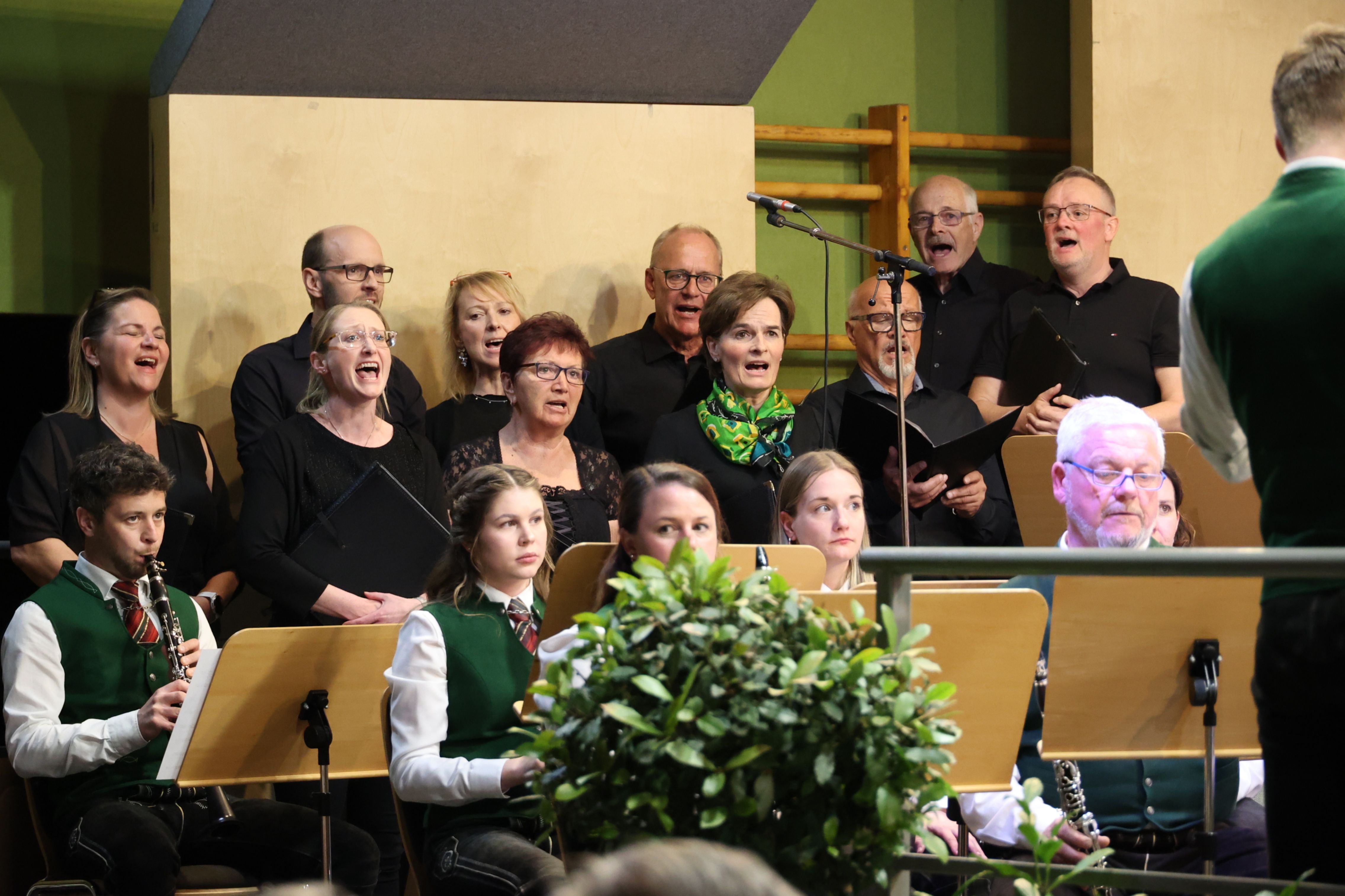 Ein Chor aus Erwachsenen und Kindern, in Schwarz gekleidet, singt in einer Kirche mit einer grünen Wand und einem hölzernen Hintergrund.