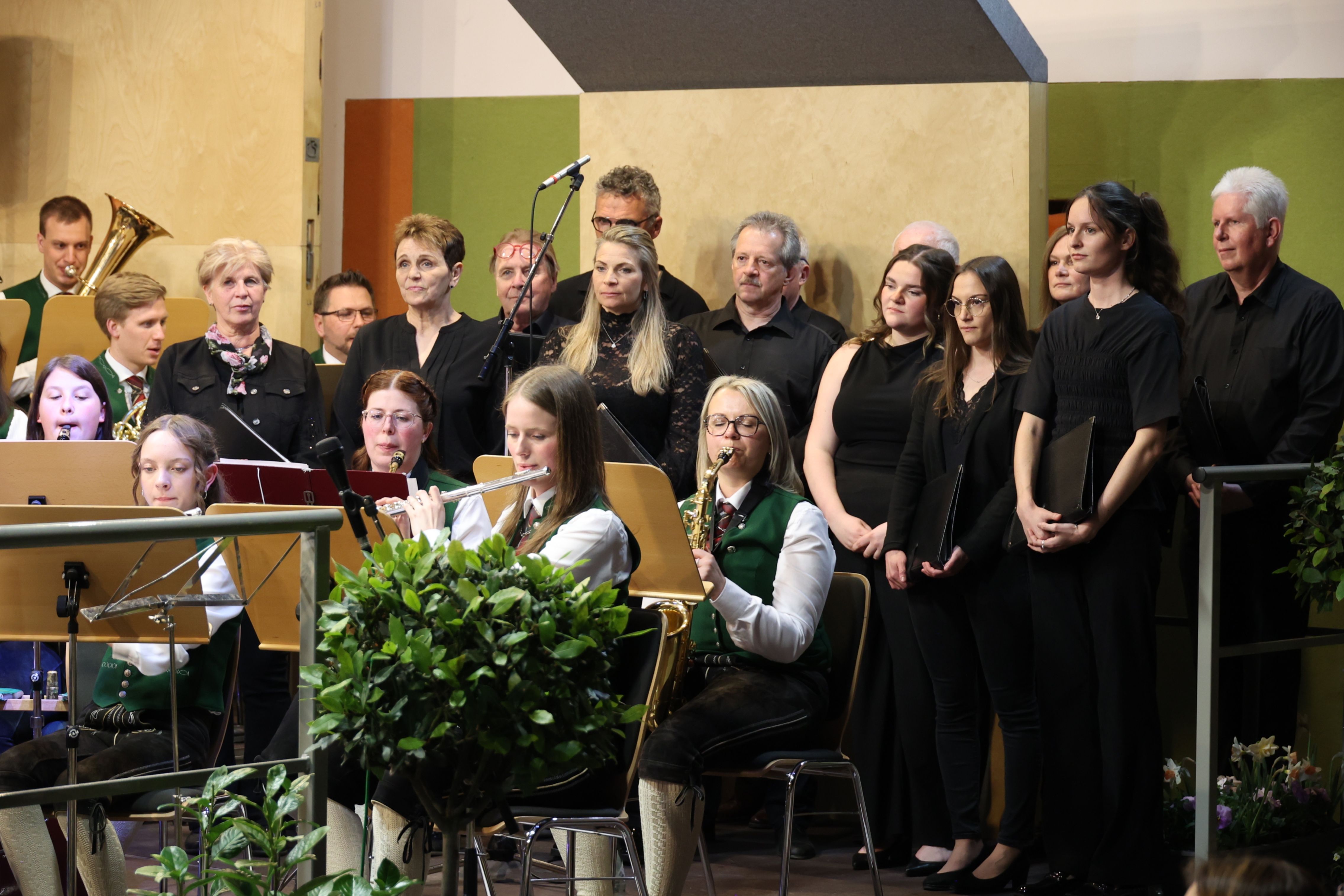 Eine Gruppe von Musikern, darunter eine Frau, die Flöte spielt, und eine andere, die Saxophon spielt, stehen in einem Raum mit einer Pflanze und einem Mikrofon.