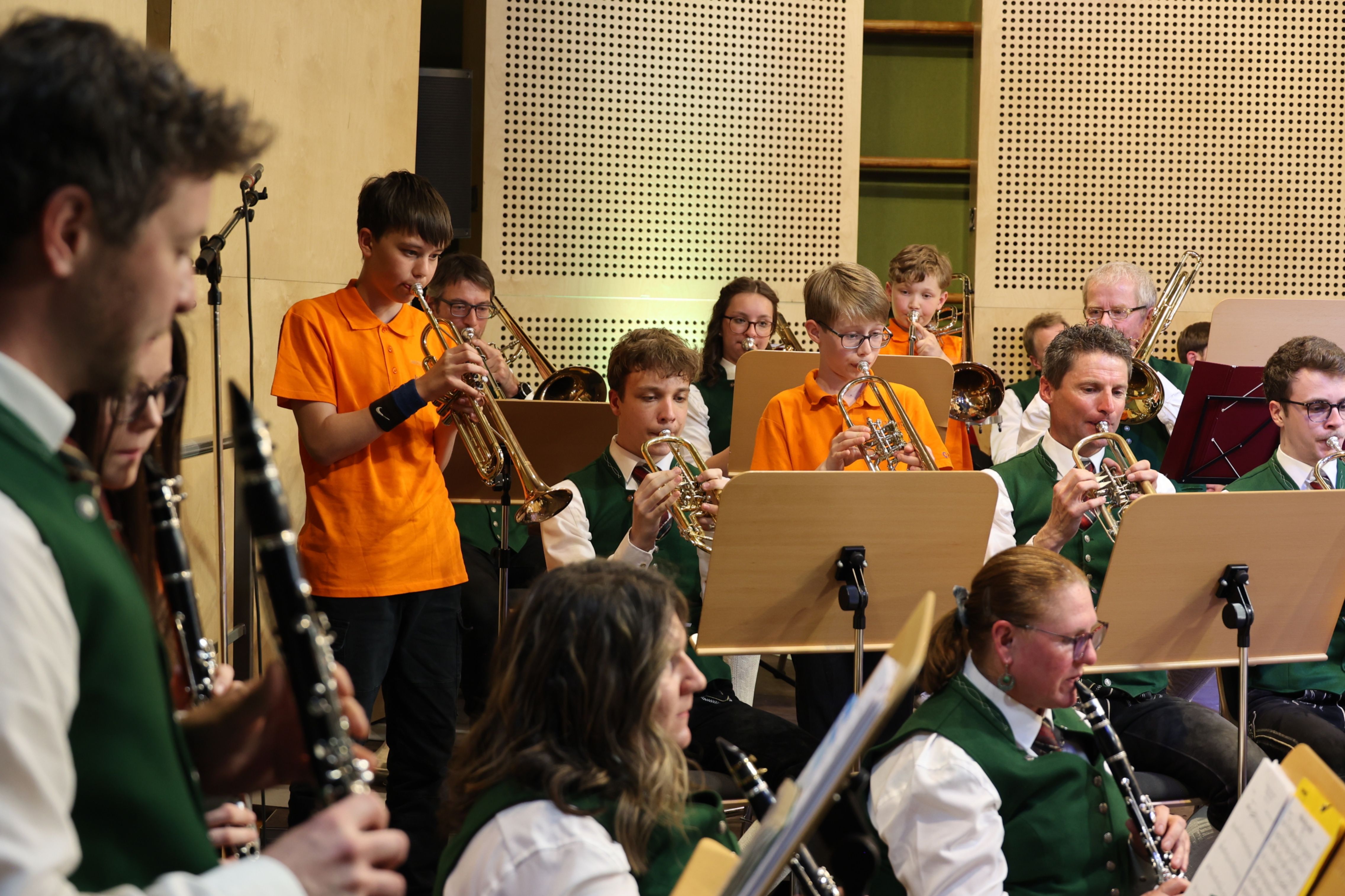 Eine Gruppe junger Musiker, darunter Jungen und Mädchen, spielt verschiedene Instrumente wie Trompeten, Posaunen und Klarinetten in einem Raum.