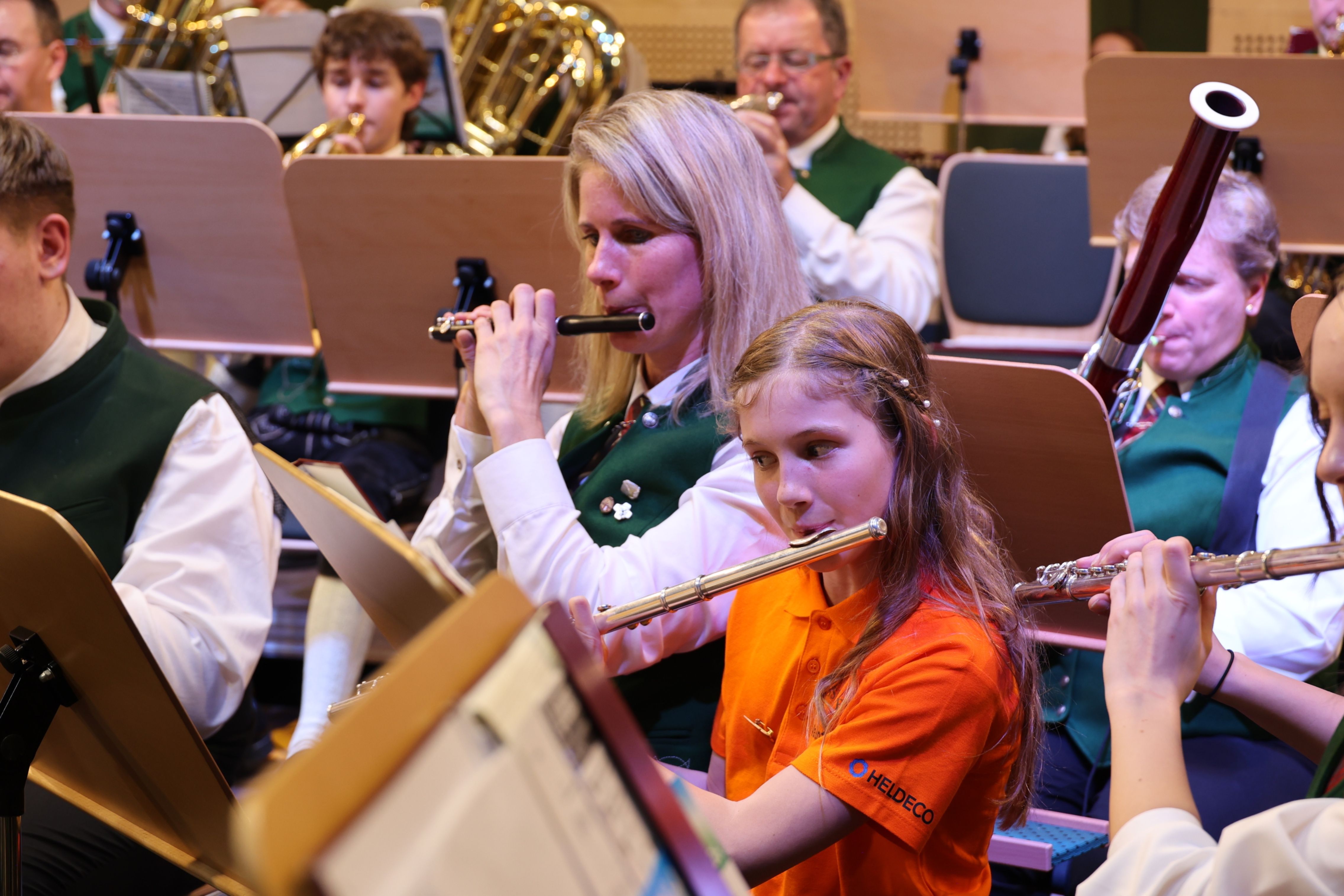 Eine Gruppe von Musikern, darunter eine Frau und ein junges Mädchen, spielen Flöten in einem Konzert. Die Frau trägt eine grüne Weste und das Mädchen ein orangefarbenes Shirt.