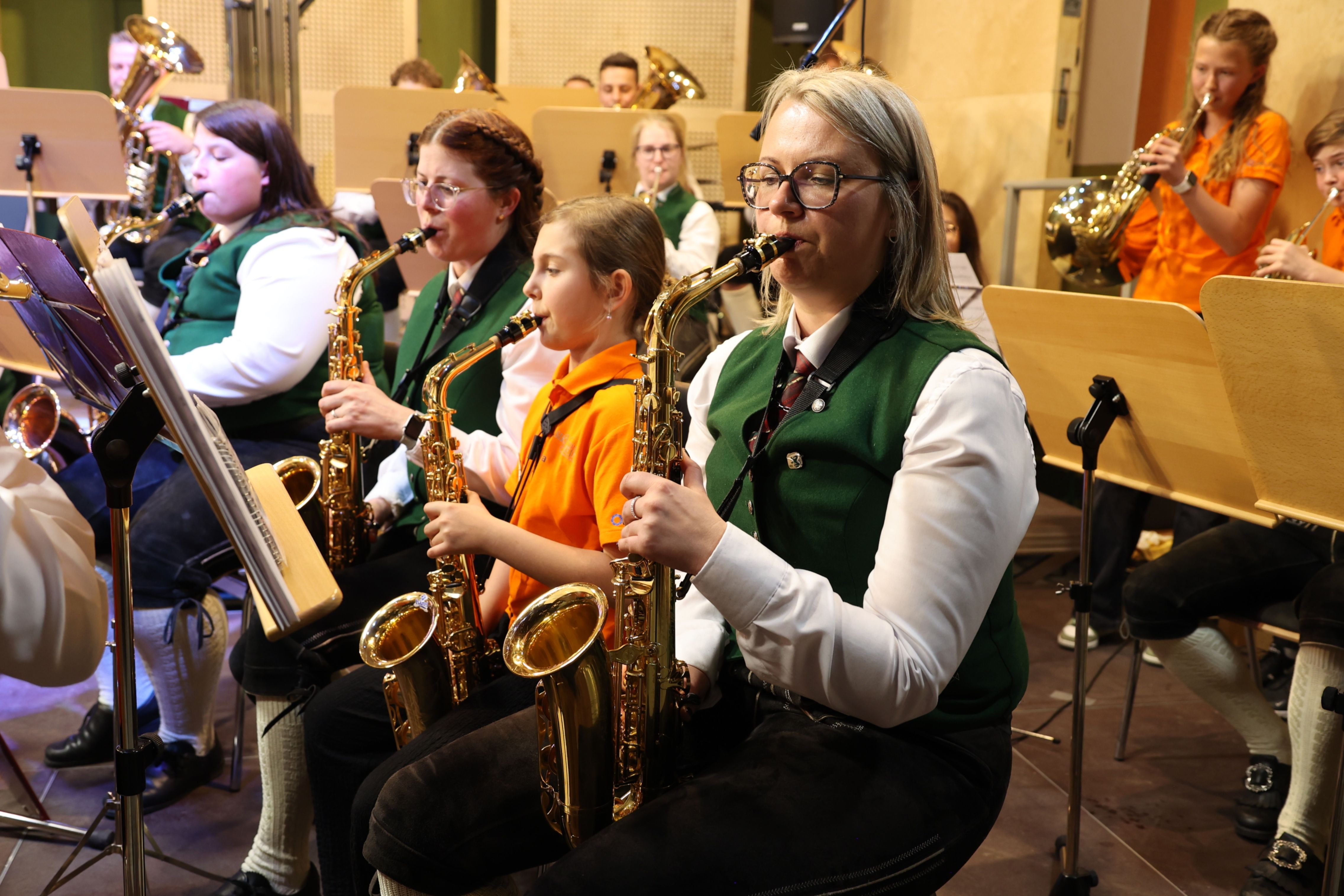 Eine Gruppe von Musikern, darunter ein junges Mädchen, spielt Saxophon in einem Musikraum. Sie tragen grüne Westen und weiße Hemden.