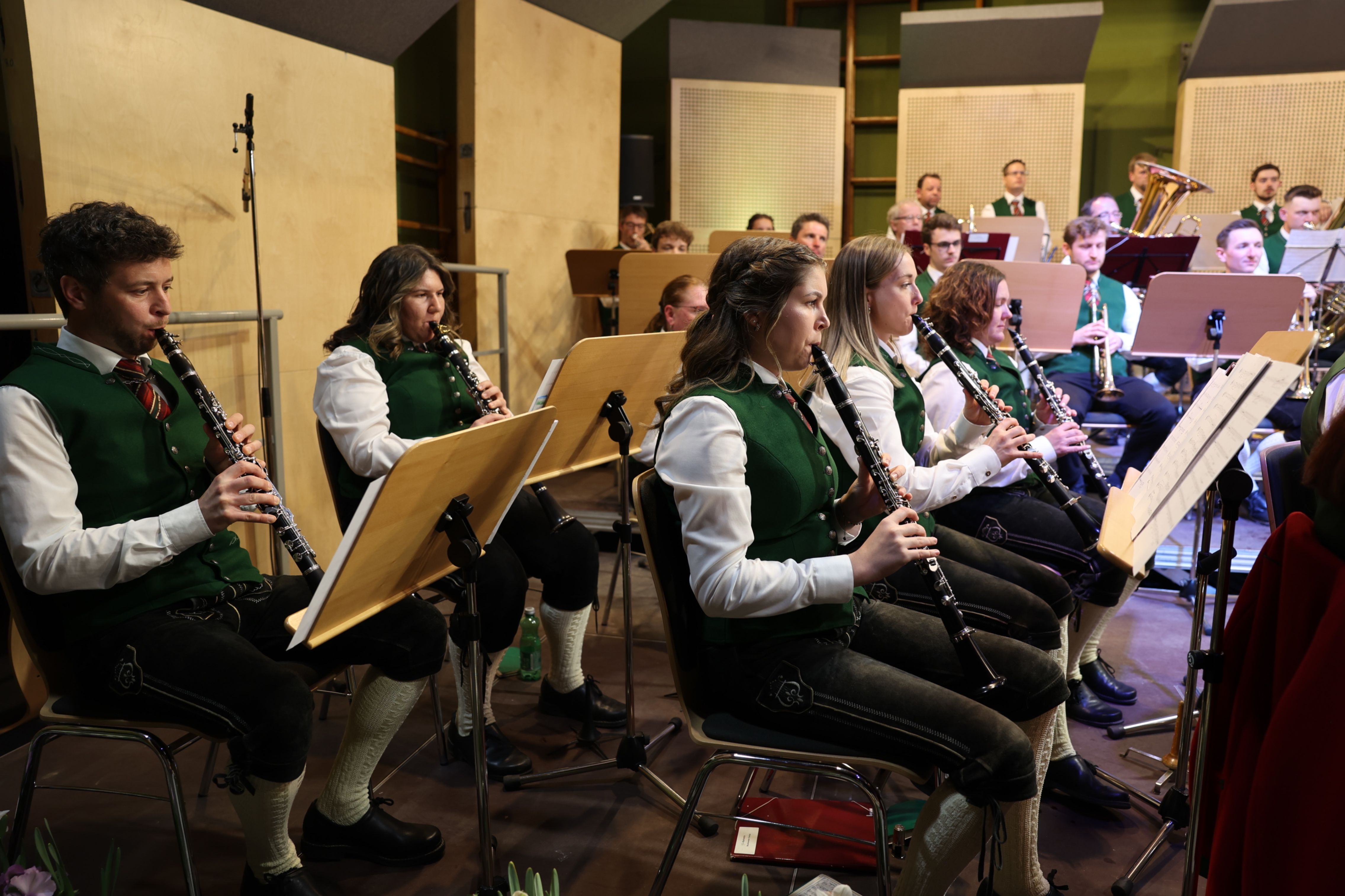 Eine Gruppe von Musikern in grünen Westen und weißen langen Ärmeln spielt Klarinetten in einem Konzertsaal. Sie sitzen auf Stühlen und haben Notenständer vor sich.