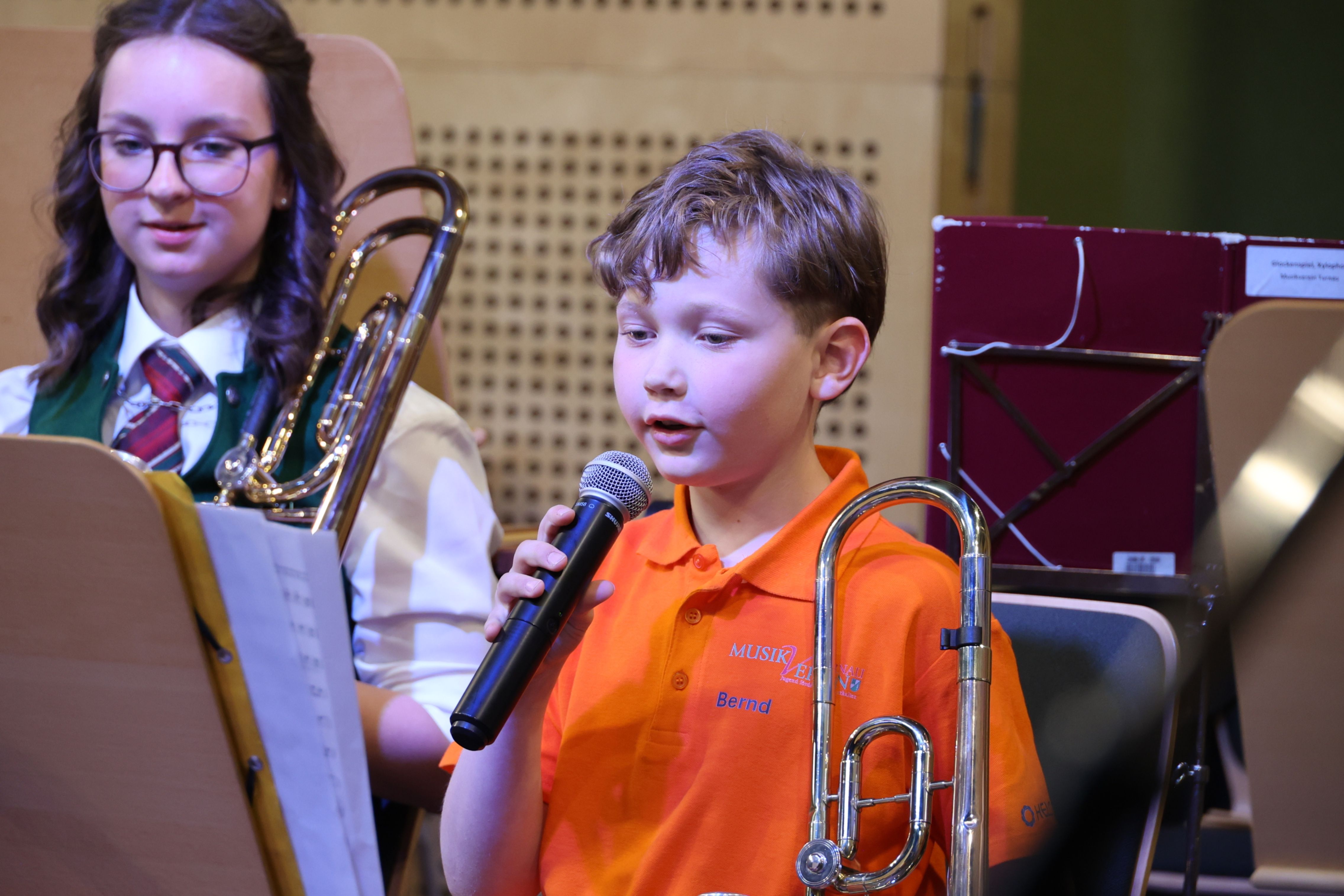 Ein junger Junge in einem orangefarbenen Shirt hält ein Mikrofon und scheint in einem Musikraum zu singen oder zu sprechen.