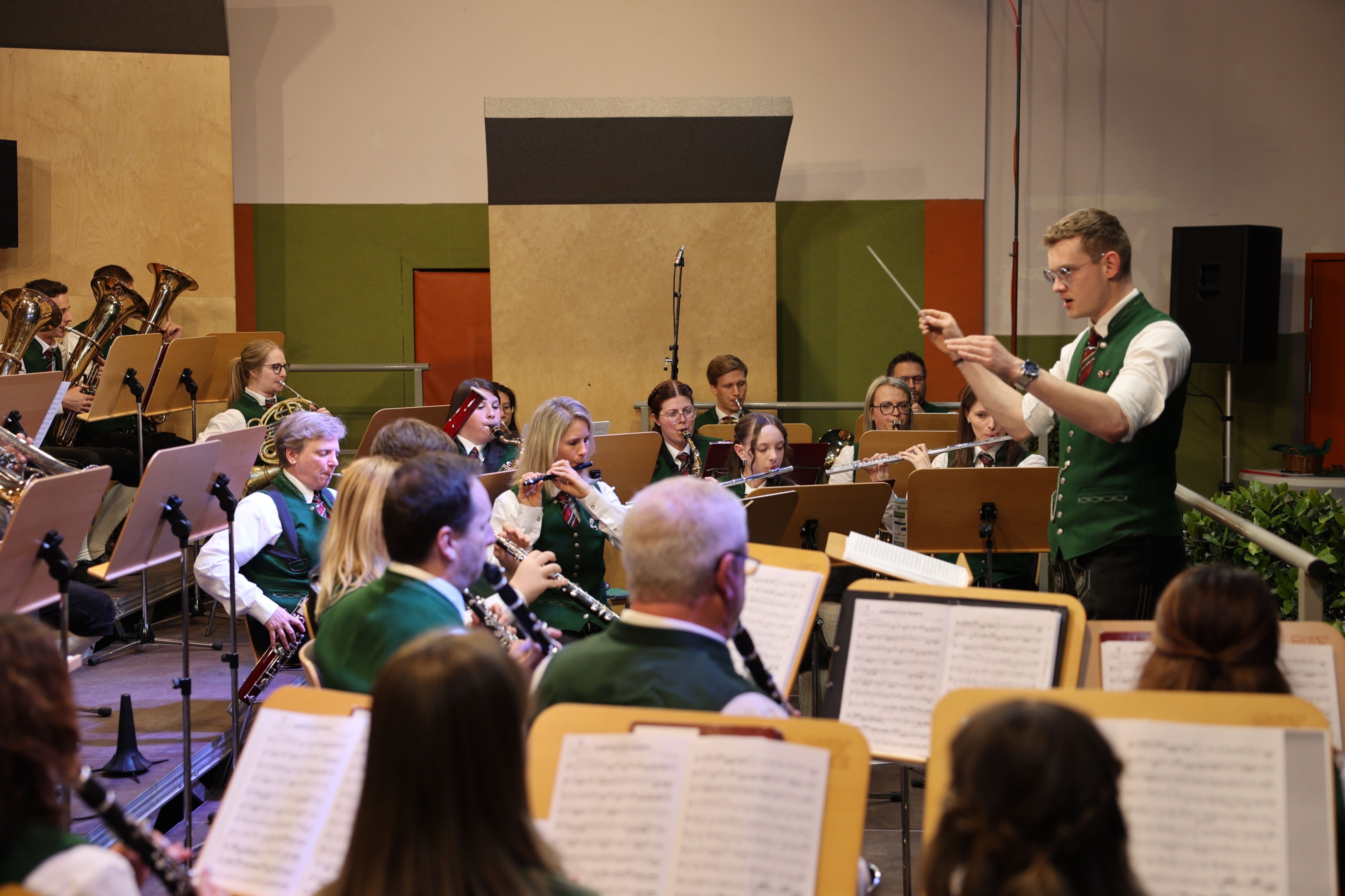 Ein Orchester spielt in einem Saal, angeführt von einem Dirigenten. Die Musiker spielen verschiedene Instrumente und das Publikum schaut zu.