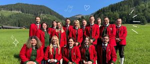 Eine Gruppe von Menschen in roter und grüner traditioneller Kleidung posiert für ein Foto auf einer Wiese mit einem Gebirgszug im Hintergrund. Die Worte 'Vorstand' und 'Musikverein Turnau' sind prominent angezeigt.