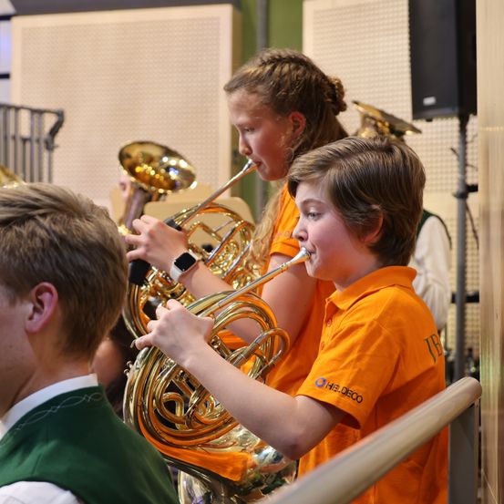 Zwei Kinder in orangefarbenen Shirts spielen Blechblasinstrumente, wahrscheinlich französische Hörner, in einem Musikraum. Ein Junge konzentriert sich auf sein Instrument, während ein Mädchen neben ihm lächelt.
