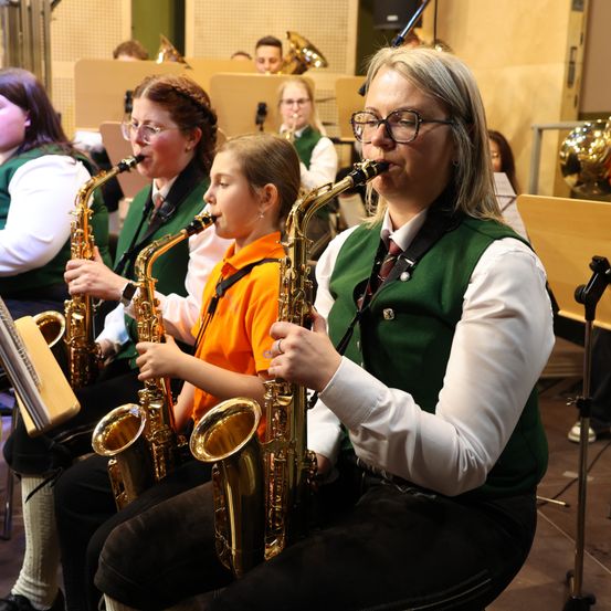Eine Gruppe von Musikern, darunter ein junges Mädchen, spielt Saxophon in einem Raum mit einer Holzwand und einem Mikrofonstativ.