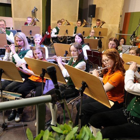 Eine Gruppe von Musikern, darunter junge Mädchen, spielt Flöten in einem gut beleuchteten Raum mit Notenständern und Instrumenten im Hintergrund.