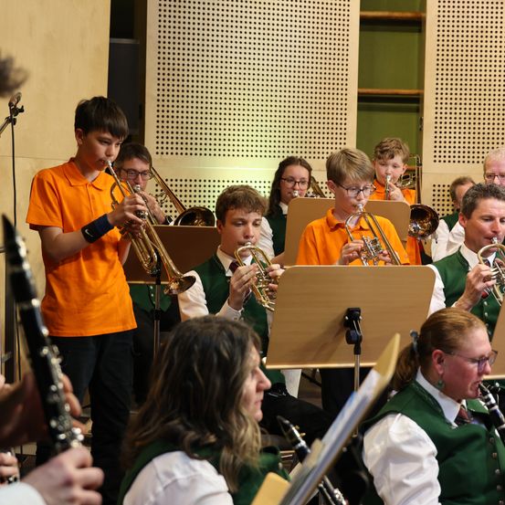 Eine Gruppe junger Musiker in grünen Westen und orangefarbenen Hemden spielt ihre Instrumente in einem Raum.