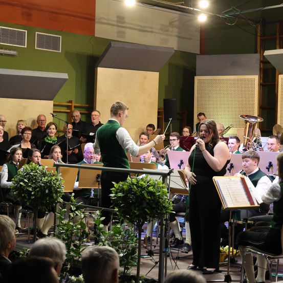 Ein Chor und ein Orchester treten auf der Bühne auf, mit einem Dirigenten und einer Sängerin, die die Aufführung leitet. Das Publikum sitzt vor der Bühne.
